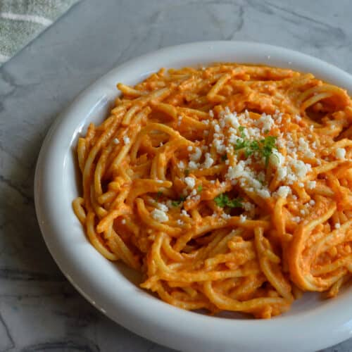 white plate of Mexican spaghetti in orange sauce topped with crumbled white cheese and herbs