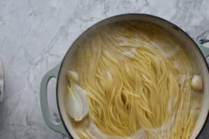 Spaghetti noodles boiling in a pot of water alongside a large piece of white onion and whole garlic cloves.