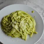 A plate of creamy green espagueti verde pasta topped with grated cotija cheese, served on a round white plate over a grey marble countertop.