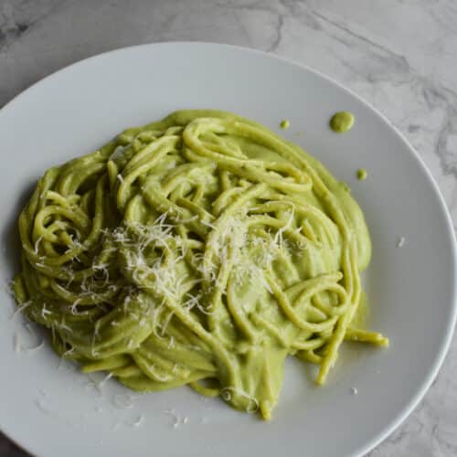 A plate of creamy green espagueti verde pasta topped with grated cotija cheese, served on a round white plate over a grey marble countertop.