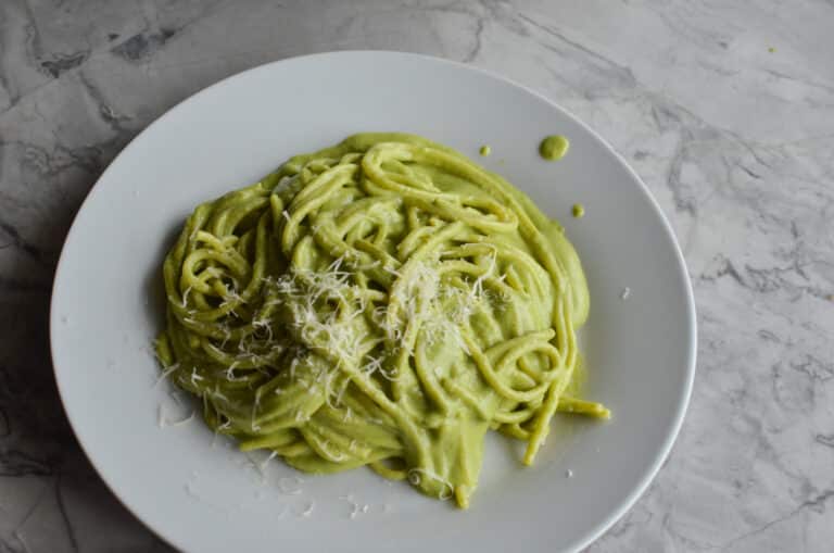 A plate of creamy green espagueti verde pasta topped with grated cotija cheese, served on a round white plate over a grey marble countertop.