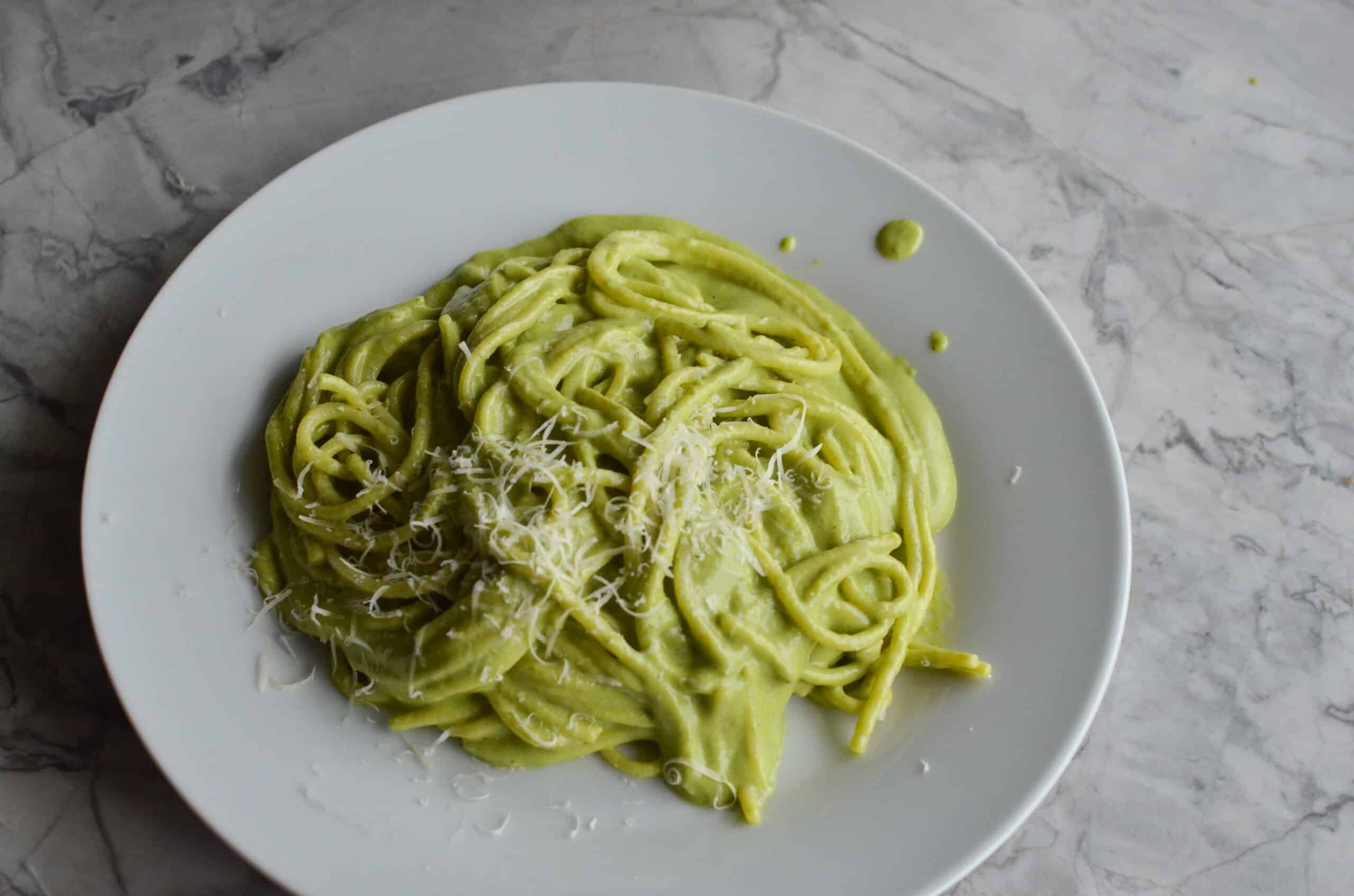 A plate of creamy green espagueti verde pasta topped with grated cotija cheese, served on a round white plate over a grey marble countertop.