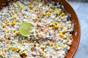 A completed Mexican street corn dip (esquites) in a brown bowl, lightly dusted with chili powder, garnished with extra cotija cheese, and topped with a fresh lime wheel in the center.