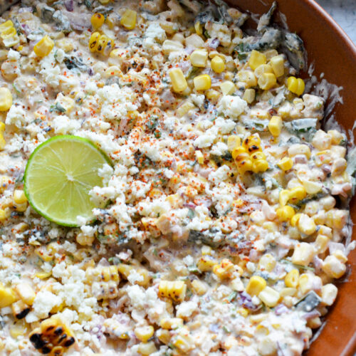 A completed Mexican street corn dip (esquites) in a brown bowl, lightly dusted with chili powder, garnished with extra cotija cheese, and topped with a fresh lime wheel in the center.