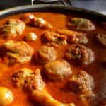 Close-up, angled view of cooked chicken drumsticks and tender potatoes bubbling in a thick, deep red tomato broth with visible herbs and spices.