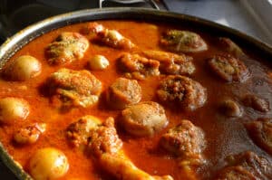 Close-up, angled view of cooked chicken drumsticks and tender potatoes bubbling in a thick, deep red tomato broth with visible herbs and spices.