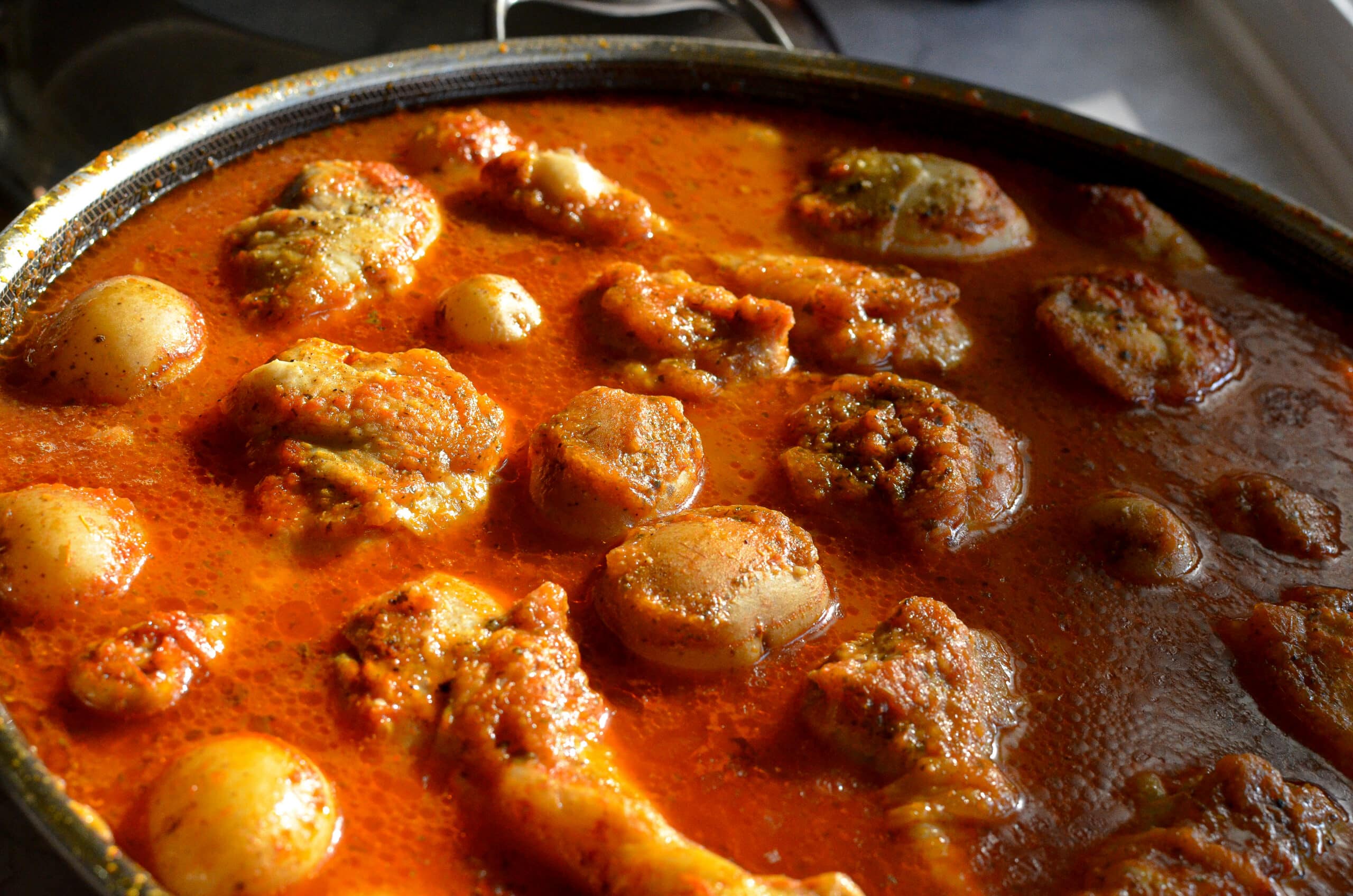 Close-up, angled view of cooked chicken drumsticks and tender potatoes bubbling in a thick, deep red tomato broth with visible herbs and spices.