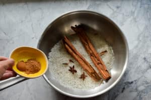 Raw white rice in a stainless steel pan with whole cinnamon sticks, star anise, and cloves, beside a yellow bowl of ground spices.