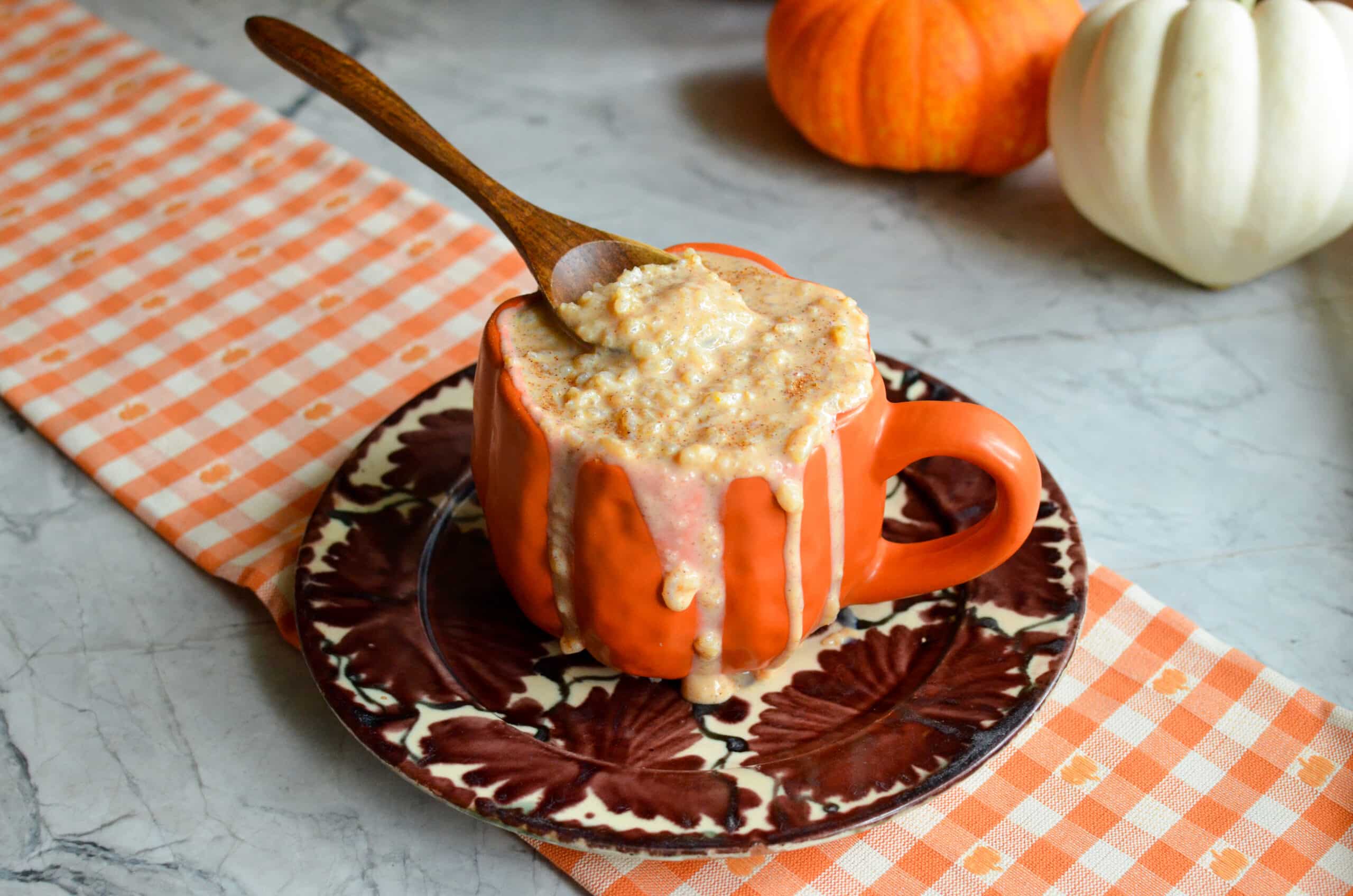 Creamy fall rice pudding overflowing an orange mug with a wooden spoon, styled with mini pumpkins.