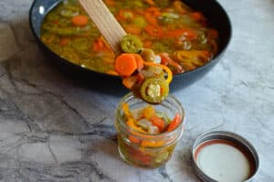 Transferring warm pickled jalapeños and carrots from a pan into a glass mason jar using a wooden spoon.