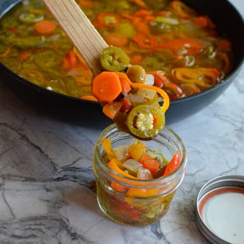 Transferring warm pickled jalapeños and carrots from a pan into a glass mason jar using a wooden spoon.