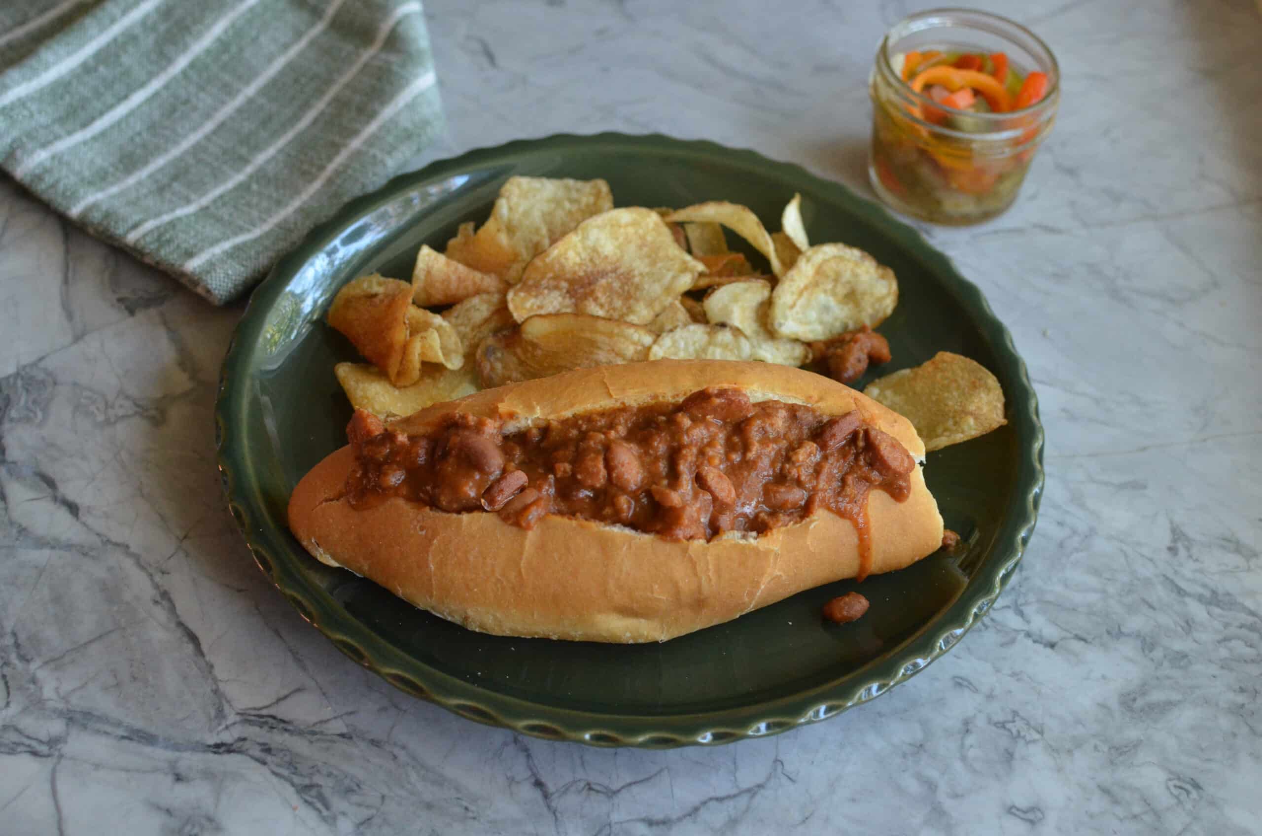 A completed Monterrey-style chili dog served on a plate with a side of crispy potato chips.