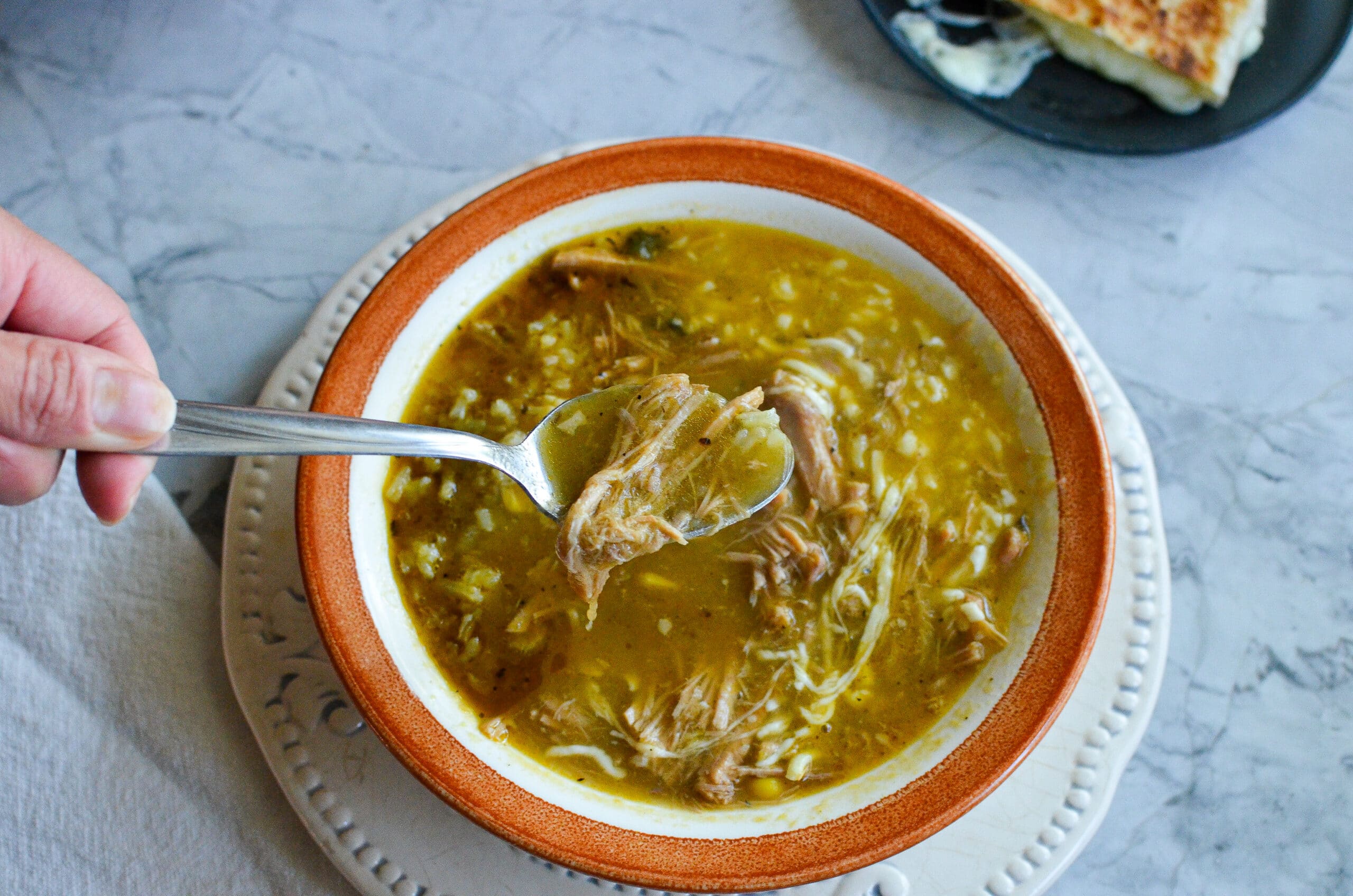 A spoon lifting tender shredded pork from a bowl of green chile stew topped with melted cheese.
