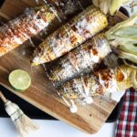 Four fully prepared Mexican street corn elotes with husks attached, arranged on a wooden cutting board with a fresh lime half and a kitchen brush nearby.