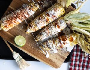 Four fully prepared Mexican street corn elotes with husks attached, arranged on a wooden cutting board with a fresh lime half and a kitchen brush nearby.