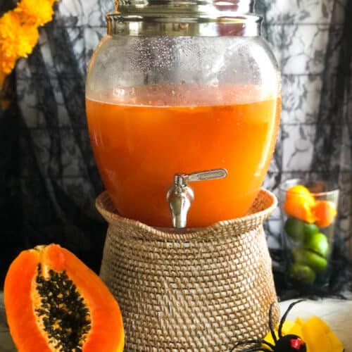Large glass drink dispenser with a silver spigot filled with vibrant orange papaya agua fresca, placed on a wicker stand next to a filled glass garnish.