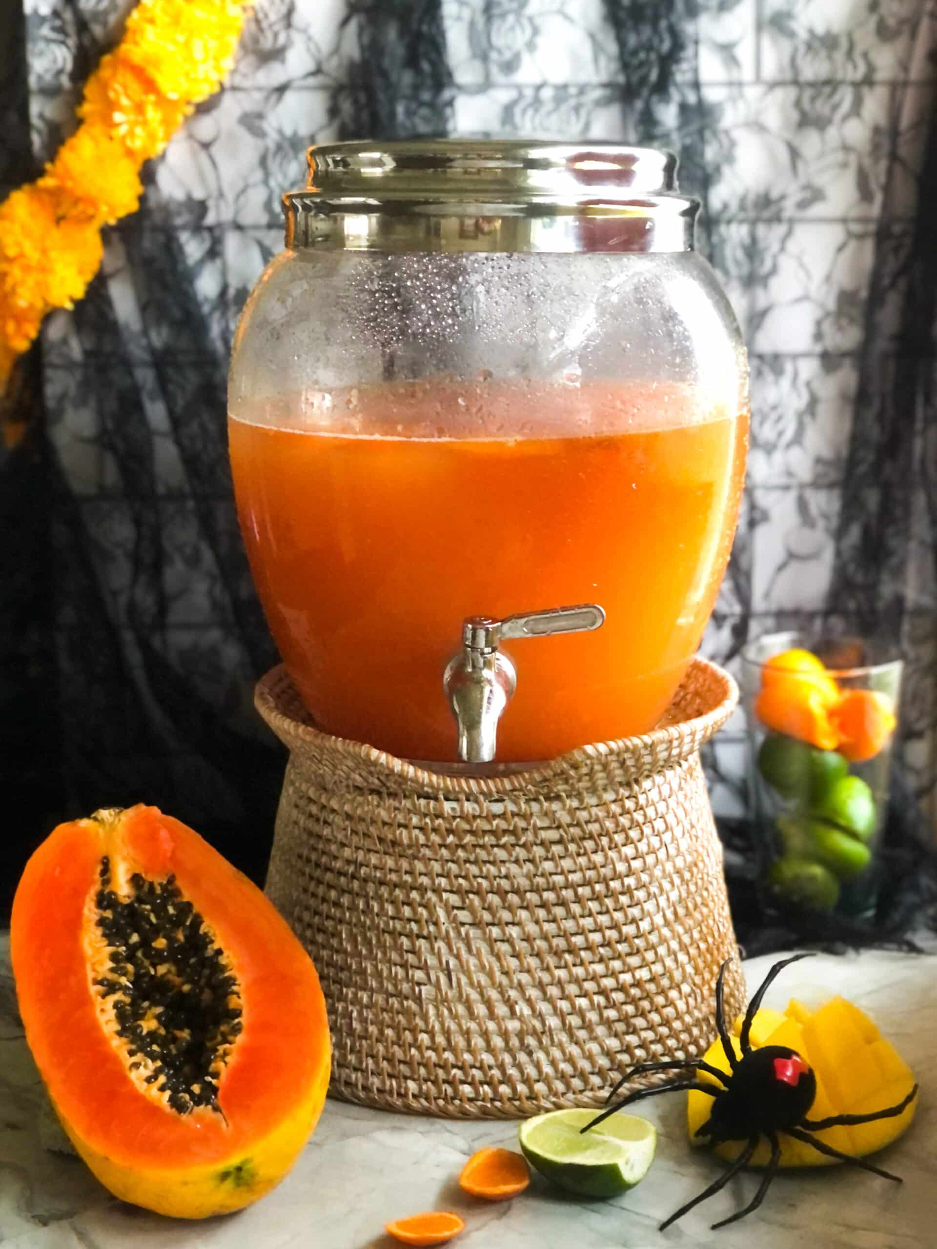 Large glass drink dispenser with a silver spigot filled with vibrant orange papaya agua fresca, placed on a wicker stand next to a filled glass garnish.
